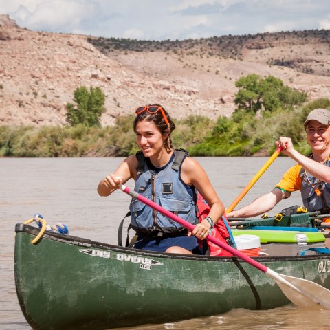 Two people paddling a canoe on a river with rocky hills in the background.