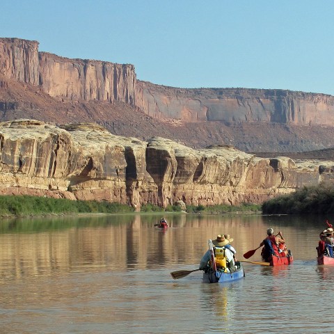 People canoeing on a river with red rock cliffs in the background.