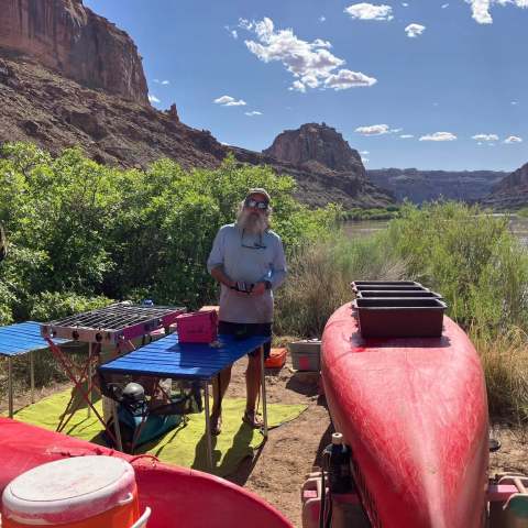 Person camping by river with red kayak and cooking station, surrounded by cliffs and greenery.