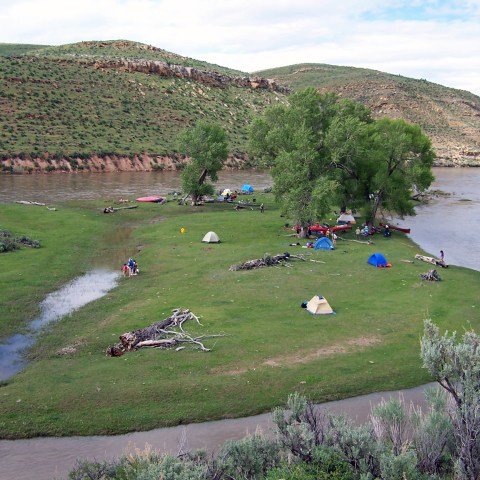 Riverside campsite with tents, trees, and people on grassy land surrounded by hills.
