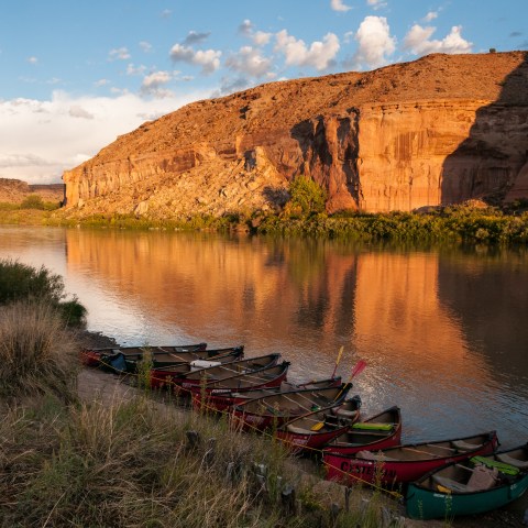 Canoes by a calm river reflecting sunlit cliffs under a blue sky.