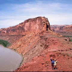 Two hikers on a red rock cliff beside a winding river and desert landscape under a clear blue sky.