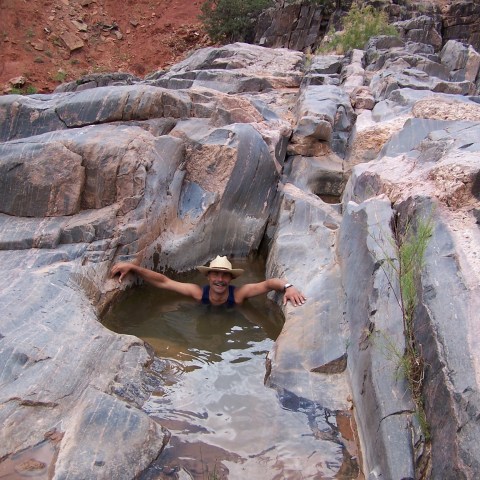Person in a hat smiling while soaking in a small natural rock pool.
