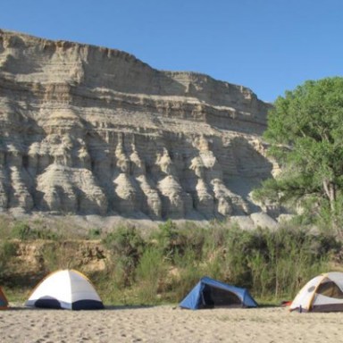 Four tents on sandy ground beneath tall rock formations and a clear blue sky.