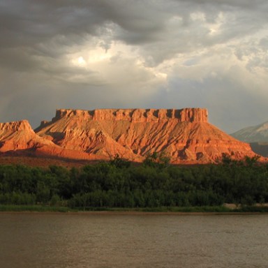 Red rock mesas under cloudy sky with river in foreground.