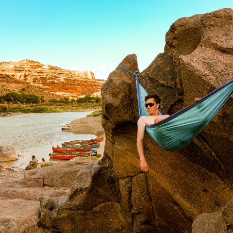Person in hammock between rocks near a river, with boats and cliffs in the background under a clear sky.