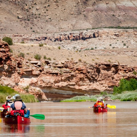 People canoeing on a river with rocky cliffs and desert landscape in the background.