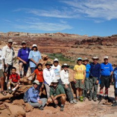 Group of people posing on rocky terrain with blue sky and canyon background.