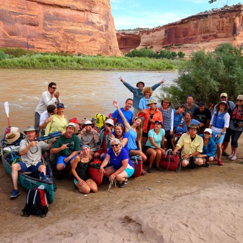 Group of people with canoes posing by a river and red rock cliffs.
