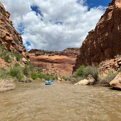 Blue rafts on a river surrounded by red rock cliffs under a partly cloudy sky.