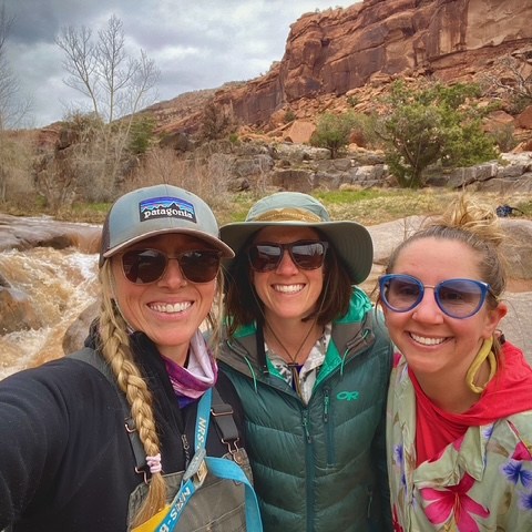 Three women smiling in outdoor attire near a rocky stream and cliffs.