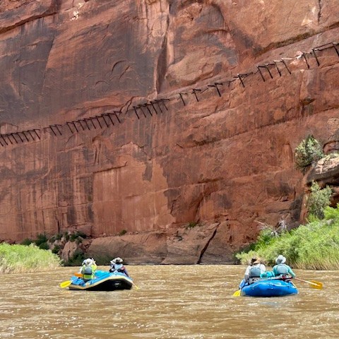 Two blue rafts with people paddling in a river, surrounded by steep red rock cliffs.