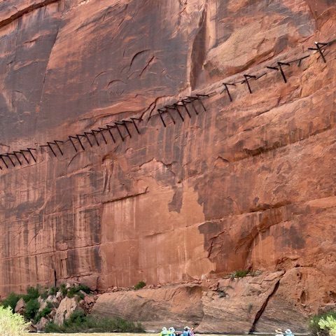 Rafts on river below steep canyon wall with wooden structure attached.