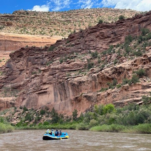 People rafting on a river with red rocky canyon walls under a blue sky.