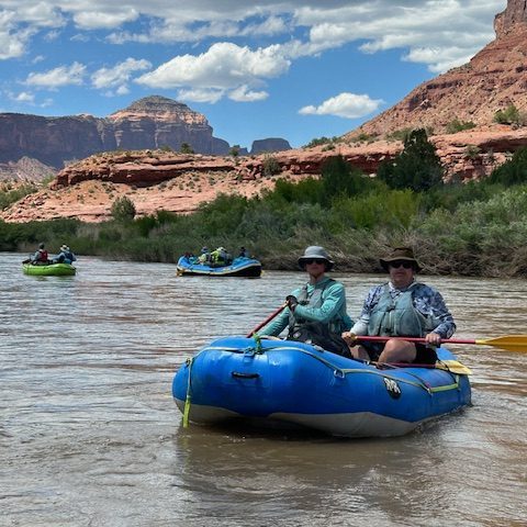 People rafting on a river with red rock cliffs and a blue sky in the background.