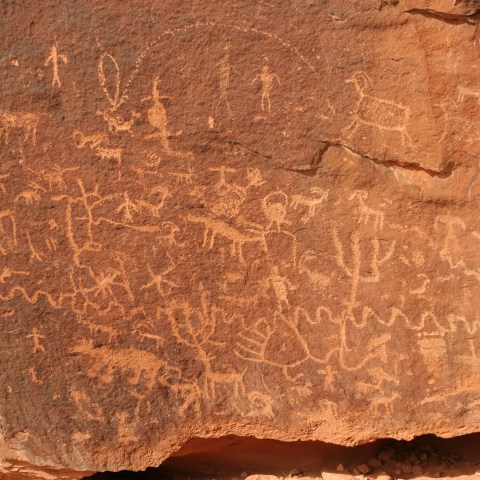 Ancient petroglyphs on a red rock surface depicting animals and human figures.