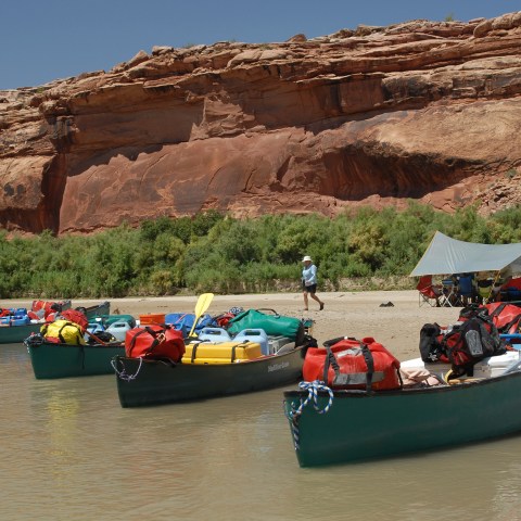 Canoes with gear docked on a riverbank with red cliffs in the background.
