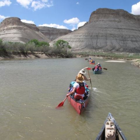 People canoeing on a river with rocky hills and trees in the background under a cloudy sky.