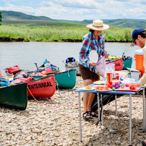 Two people prepare food on a foldable table by a riverbank with canoes and gear in the background.