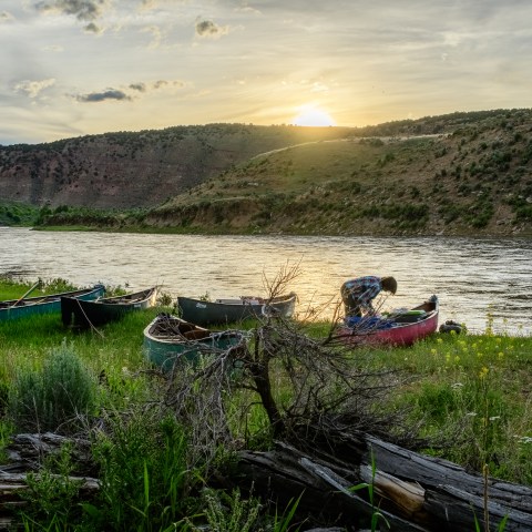Canoes on a grassy riverbank at sunset with hills in the background.