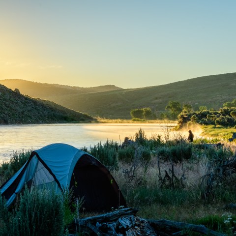 Camping tents by a river at sunrise, with mist and hills in the background.