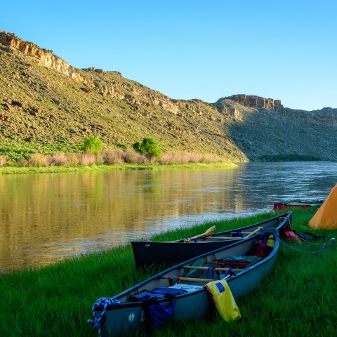 Canoes and a tent by a river with a rocky hill in the background under a clear blue sky.