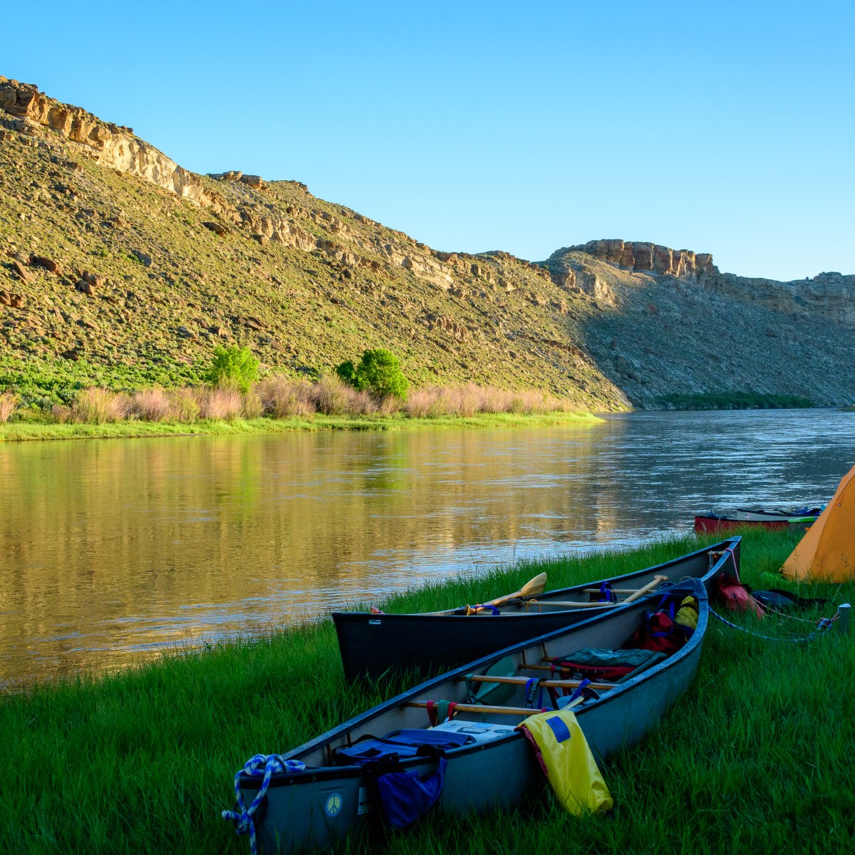 Canoes and a tent by a river with a rocky hill in the background under a clear blue sky.
