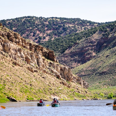People canoeing on a river surrounded by rocky hills and trees under a clear sky.