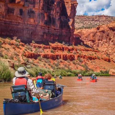 Canoeists paddle through a river canyon with red rock cliffs under a blue sky.