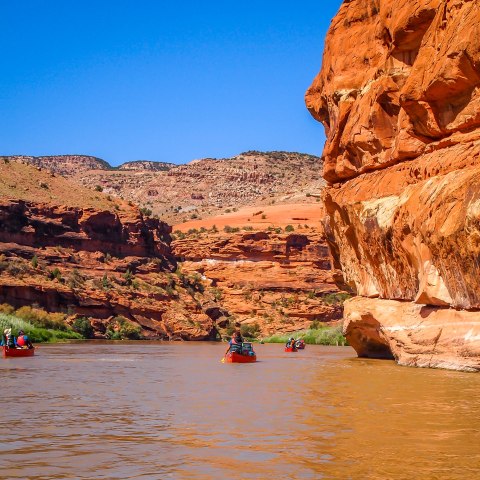 Canoes on a river surrounded by red rock cliffs under a clear blue sky.