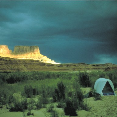 Campsite with tents in desert landscape, dramatic lighting on distant cliffs under stormy sky.