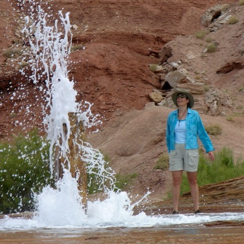 Person with hat stands near geyser erupting in rocky landscape.