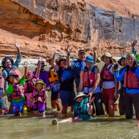 Group of people in life jackets cheering in a river with rocky cliffs in the background.