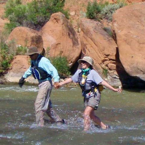 Two people holding hands, wading through a shallow river near large rocks.
