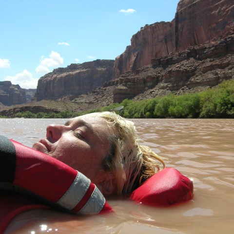 Person in red life jacket floating in river with canyon background.