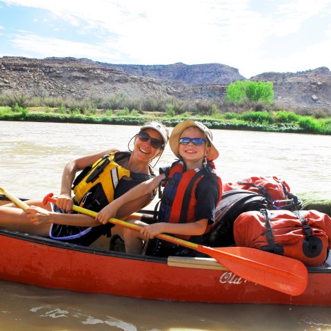 Two people in a red canoe on a river, wearing life jackets and smiling under a sunny sky.