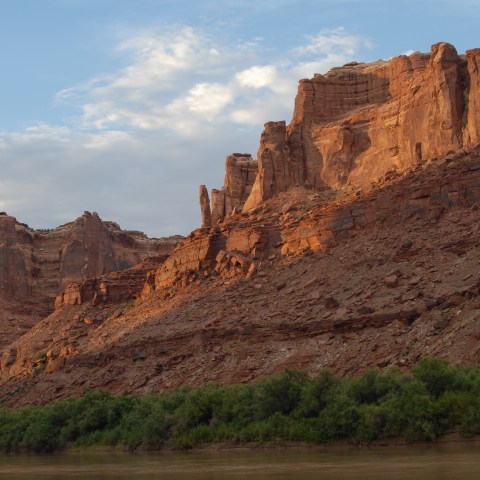 Red rock cliffs overlook a river with green bushes under a blue sky with clouds.