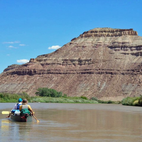 Two people in a canoe paddle on a river with a large rocky hill in the background.