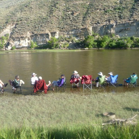 People in chairs relax by a riverbank with a rocky hillside in the background.