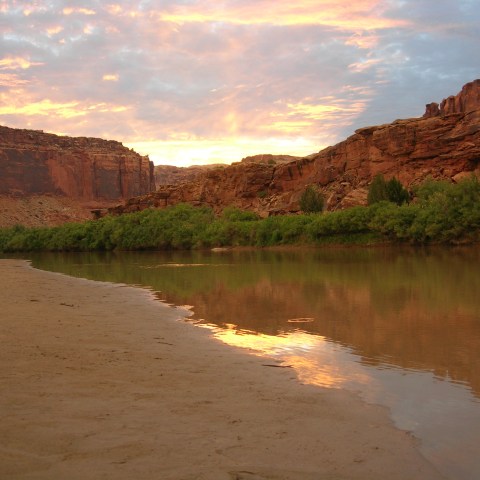 Tranquil river reflecting colorful sunset with rocky cliffs and greenery along the bank.