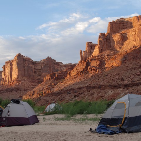 Tents pitched on a sandy ground with towering red rock formations under a partly cloudy sky.