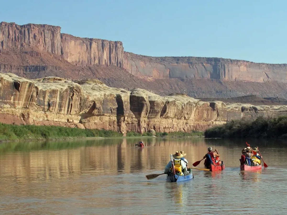 People in canoes paddle on a river with red rock cliffs in the background.